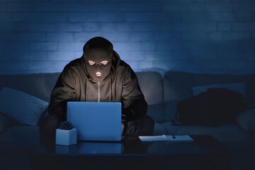 Fototapete - Masked hacker wearing black balaclava sitting at a desk on couch, downloading private information off computer at night