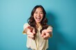 © velikiyzayats - Cheerful Asian Young Woman in Yellow Blouse Smiling and Pointing Playfully Against a Vibrant Teal Background in a Bright, Energetic Setting