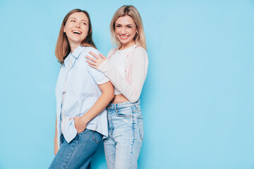 Wall Mural - Two young beautiful smiling brunette and blond hipster female in trendy summer clothes. Lovely women posing on blue background in studio. Positive models having fun. Cheerful and happy. isolated