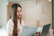 © Ekkasit A Siam - Asian woman holding a document and reading attentively while working on a laptop in a bedroom. Focused expression suggests serious work, remote job, or online education in a home-based setting.