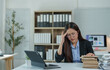 © crizzystudio - Stressed asian businesswoman touching her forehead while experiencing a headache, surrounded by documents and a digital tablet at her cluttered office desk, reflecting tension and fatigue