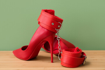 Wall Mural - Red handcuffs from sex shop and stylish female high-heeled shoes on table near green wall in room, closeup