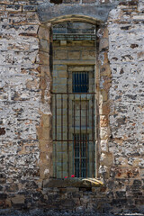 A weathered stone wall frames a barred window, hinting at forgotten stories and history, with faint blossoms peeking through, adding a touch of color and life.