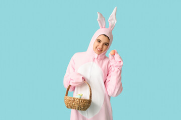 Wall Mural - Beautiful young woman in bunny costume holding wicker basket with Easter cake and eggs on blue background