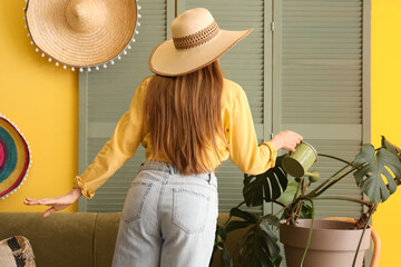 Wall Mural - Young woman in hat with houseplants in yellow living room, back view