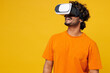 © ViDi Studio - Young gambling happy cheerful fun cool Indian man he wears orange t-shirt casual clothes watching in vr headset pc gadget isolated on plain yellow wall background studio portrait. Lifestyle concept.