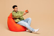 © ViDi Studio - Full body young man of African American ethnicity wear green casual clothes sit in bag chair use play racing app on mobile cell phone hold gadget for pc video games isolated on plain beige background.