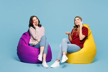 Canvas Print - Full body young couple two friends women wearing red casual clothes together sit in bag chair talk speak on mobile cell phone isolated on pastel plain light blue background studio. Lifestyle concept.