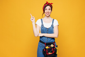 Sticker - Young fun employee laborer handyman woman wear overalls belt point index finger aside on empty area isolated on plain yellow background. Instruments for renovation apartment room. Repair home concept.