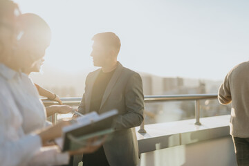 Wall Mural - Group of business people standing on a high-rise balcony, discussing ideas and project solutions. The setting sun creates a warm atmosphere of teamwork and innovation.