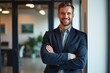 © SimpleDesignStudio - Young Smiling Caucasian Male Professional in Smart Suit Standing with Arms Crossed in Modern Office Environment, Exuding Confidence and Approachability