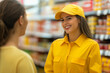 © Regina Gafarova - A smiling young female sales consultant in a yellow uniform talks to a customer in front of a supermarket.