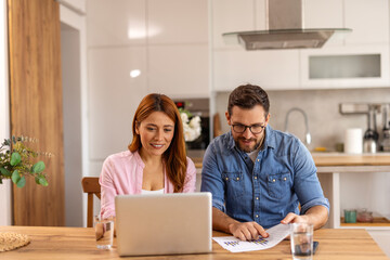 Couple is sitting at a table, focused on a laptop screen. They be discussing or working together, highlighting themes of collaboration, technology, and modern lifestyle in a home office setting