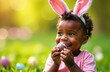 © Yuliya - Happy black boy with bunny ears on his head eats a chocolate egg in the yard of the house. Children's Easter hunt.