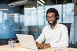 © Liubomir - Young man wearing headphones, glasses, and a white shirt works on his laptop in an office setting, smiling at the camera.