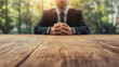 © Michael - Businessman sitting with clasped hands at wooden table in a green park, focused on the table