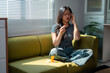 © PaeGAG - Young woman suffering from headache, holding pills and touching her head while sitting on sofa at home