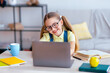 © Prostock-studio - Distance Education. Smiling female pupil in eyeglasses sitting at desk, using laptop and typing on keyboard, doing homework, watching tutorial, lecture or webinar, studying online at home. E-learning