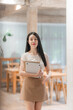 © Kritdanai - Confident Woman in Cafe: A poised young woman, with long dark hair, stands confidently in a modern cafe, holding a tablet, exuding a sense of professionalism and approachability.