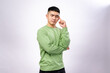 © Daniel - An Asian man in a green sweatshirt poses thoughtfully with a finger on his temple and a serious expression. He stands against a plain white background, conveying deep thinking, focus