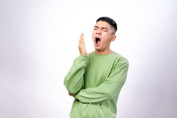 An Asian man wearing a green sweatshirt stands against a white background, yawning with his hand covering his mouth. He has his arms crossed, expressing tiredness or boredom