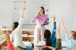© Home-stock - Cheerful teacher playing with pupils, students raising their hands up during lesson in classroom of private elementary school