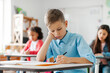 © Home-stock - Focused schoolboy leaning over his copybook, writing test in classroom with classmates sitting on background