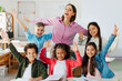 © Home-stock - Excited teacher and group of diverse school classmates raising arms and smiling, sitting in classroom, celebrating the last day of school