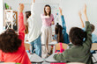 © Home-stock - Young teacher asking kids, pupils sitting with raised hands want to answer, classroom interior. Woman pointing at pupil and smiling