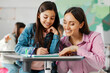 © Home-stock - Friendly teacher helping girl with homework or exam, schoolgirl writing in notebook, sitting at desk with classmates on background