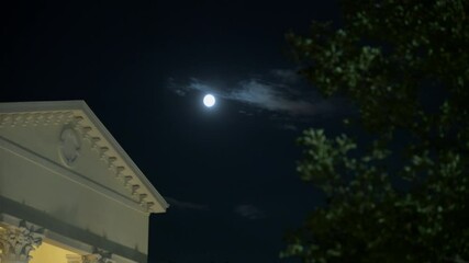 Wall Mural - The moon shines over the white columned house in the evening with a full moon in the sky. The dark sky above has a visible moon with soft glow. Tree branches frame the top of the image. The atmosphere