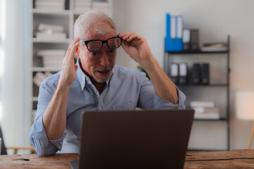 Surprised senior manager with grey hair and beard, wearing blue shirt, sitting at desk and looking at laptop while adjusting glasses in office during daytime