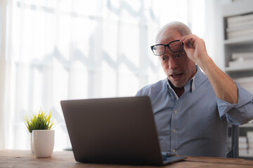 Senior businessman working from home looking surprised at his laptop raising his glasses in his modern bright office with plants and bookshelves in the background