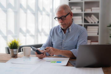 Focused mature man using calculator and analyzing documents while working with laptop computer at home, sitting at wooden table with cup of coffee and plant