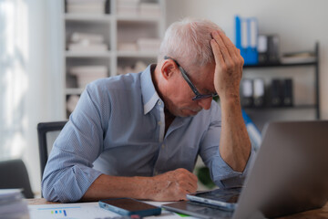 Senior businessman sitting at his office desk holding his head with his hand, showing concern and suffering from headache or migraine, having problems at work or from overworking