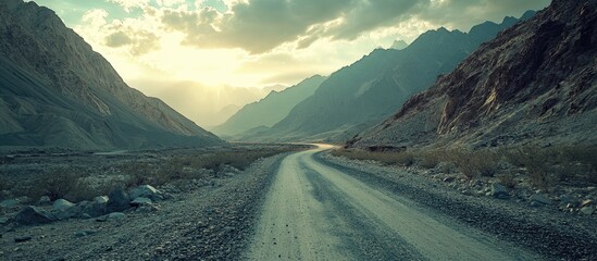 Fototapete - Serene mountain landscape with a winding dirt road in the foreground, soft sunlight filtering through clouds, rugged peaks in the background.