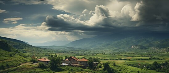 Poster - Dramatic landscape featuring a rustic farmhouse in the foreground surrounded by lush green fields under a moody, cloudy sky with distant mountains.