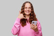 © sementsova321 - A cheerful young woman with curly brown hair is holding a smartphone and making a call gesture with her other hand, dressed in a cozy pink sweater and standing against a neutral gray background