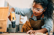 © sorapop - Carpenter woman using a power drill to secure a piece of wood in a workshop, focusing on precision in furniture making. Surrounded by tools and wood pieces, highlighting skill and craftsmanship