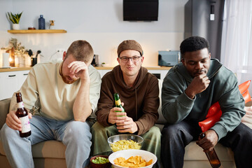 Front view portrait of three male friends sitting on couch at home and watching TV with mixed emotions