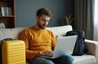 © Viktor - Young man planning vacation using laptop to book hotel room online. Male tourist wearing sweater sits on sofa browsing rental holiday accommodation website, next to yellow suitcase and black rucksack.