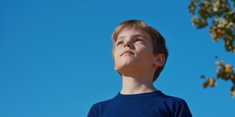 Wall Mural - A young boy is standing outside on a sunny day, looking up at the sky. The boy is wearing a blue shirt and he is enjoying the beautiful weather