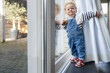 © Connect Images - Toddler in overalls standing by a glass door to the outside with a curious expression and a snack in his hand, Netherlands