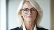 © Katerina Bond - Confident mature business Caucasian woman with blond hair and glasses, wearing a suit, looking directly at the camera with a neutral expression, in a professional indoor setting.