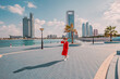 © EdNurg - Female tourist wearing red dress walking on promenade in Abu Dhabi and talking on the phone with modern skyscrapers in the background