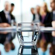 © UraiwanT - A clear glass of water sits on a reflective surface in front of a group of people engaged in a meeting.