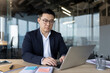 © Tetiana - A serious young Asian man in a business suit is sitting in the office at a desk and is working on a laptop with concentration