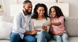© Prostock-studio - People And Technology. Portrait of happy African American family holding and using mobile phone sitting on the couch at home. People spending time together, sharing social media, watching funny video