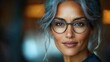 © imagineRbc - A woman with elegant grey hair and stylish glasses looks directly at the viewer. The warm lighting of a cozy space enhances her confident expression in the early evening