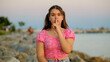 © HI Pictures - A somewhat chubby teenage girl sitting on the rocks by the seaside on a sunny summer day, looking at the camera and making a 'shush' gesture with her hand